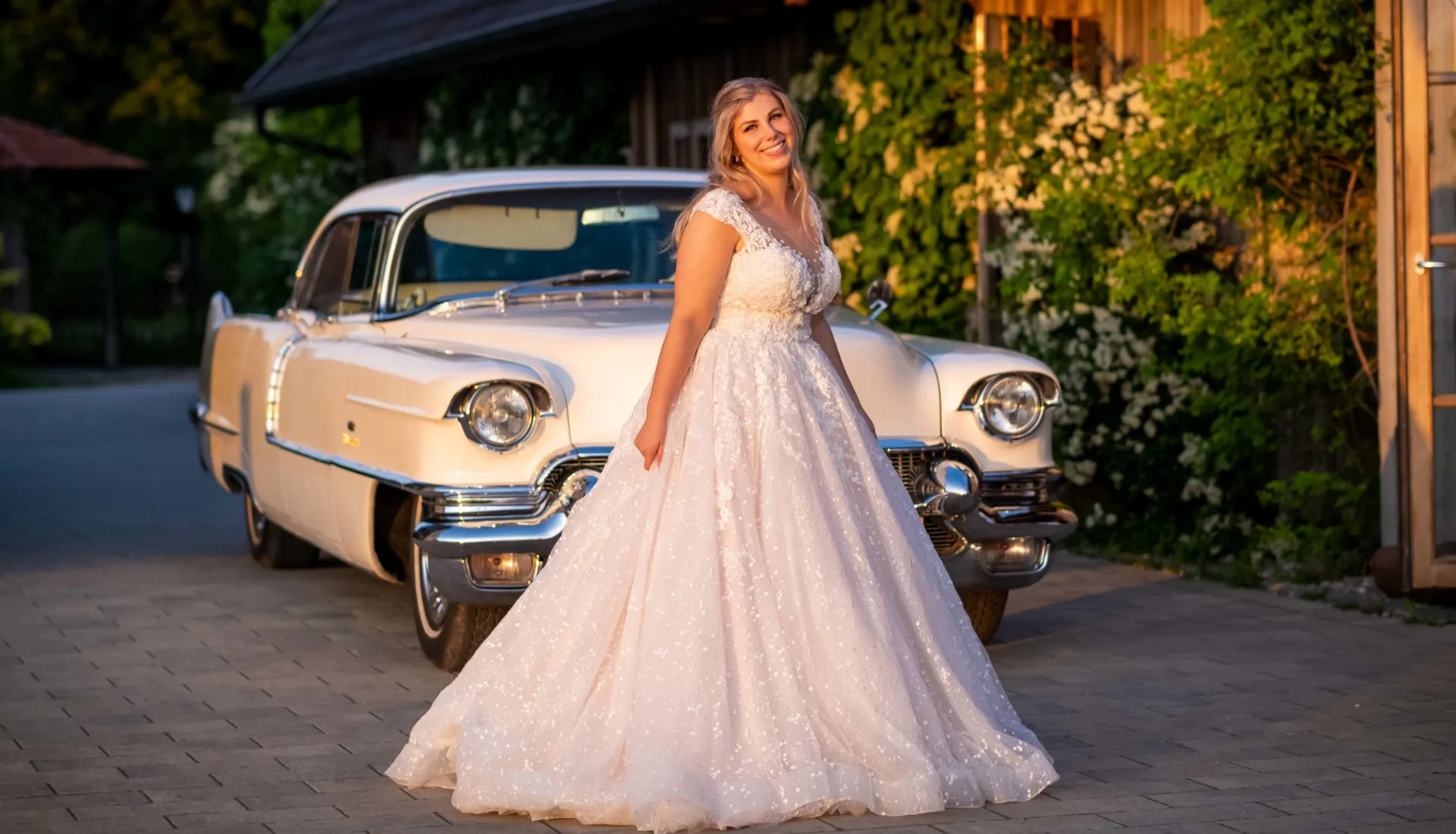 Braut im Hochzeitskleid vor einem Vintage-Auto bei einer Hochzeit im Bayerischen Oberland